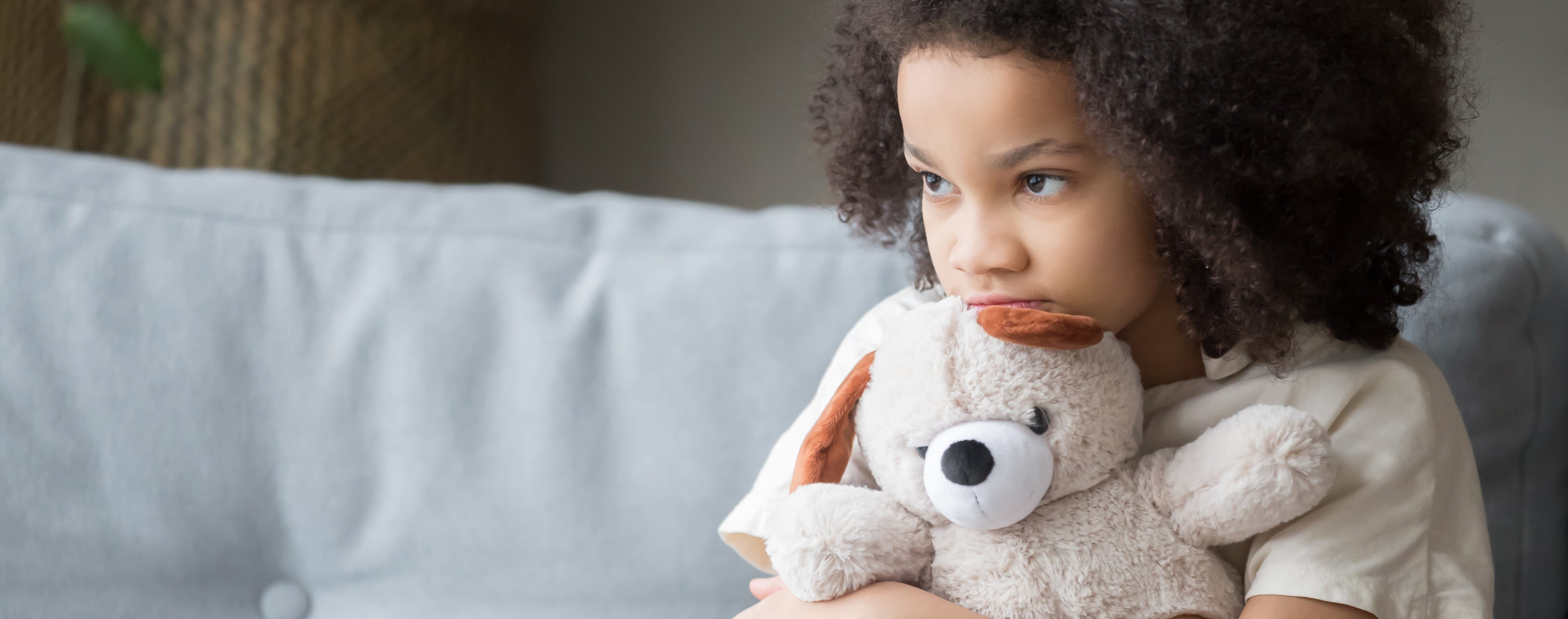 Barn med nalle Sorgset barn sitter i soffa med nalle i knät