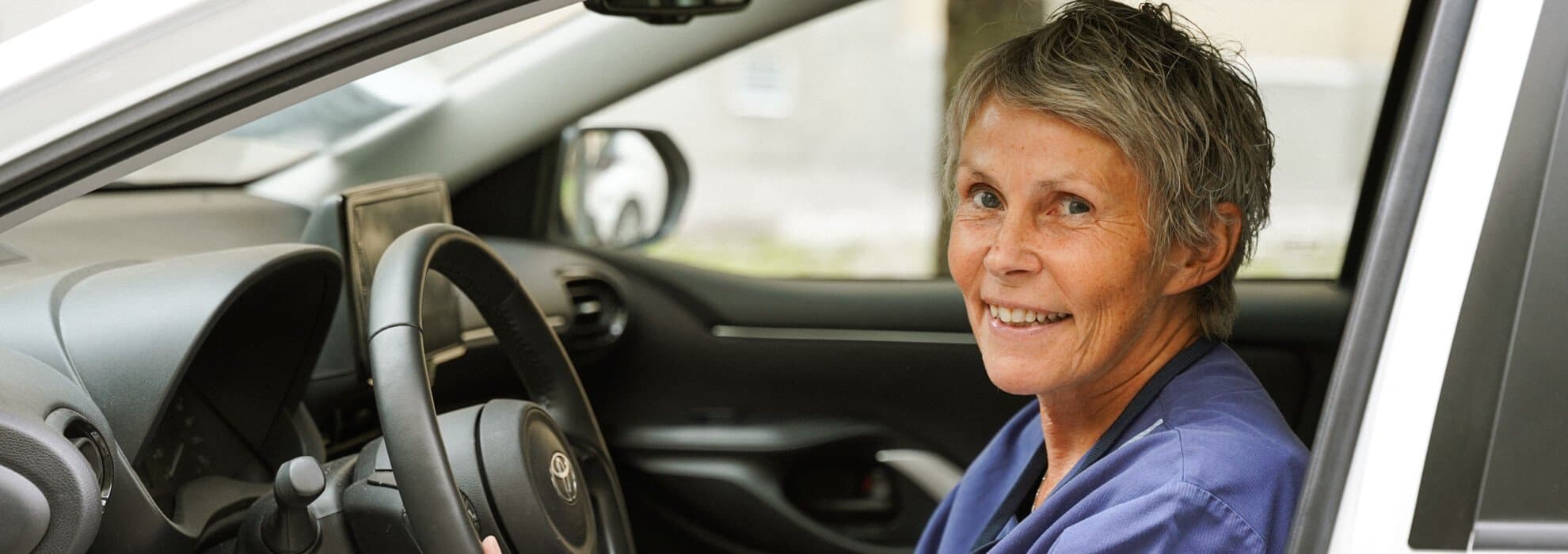 En sjuksköterska sitter i en bil med öppen dörr och tittar ut leende