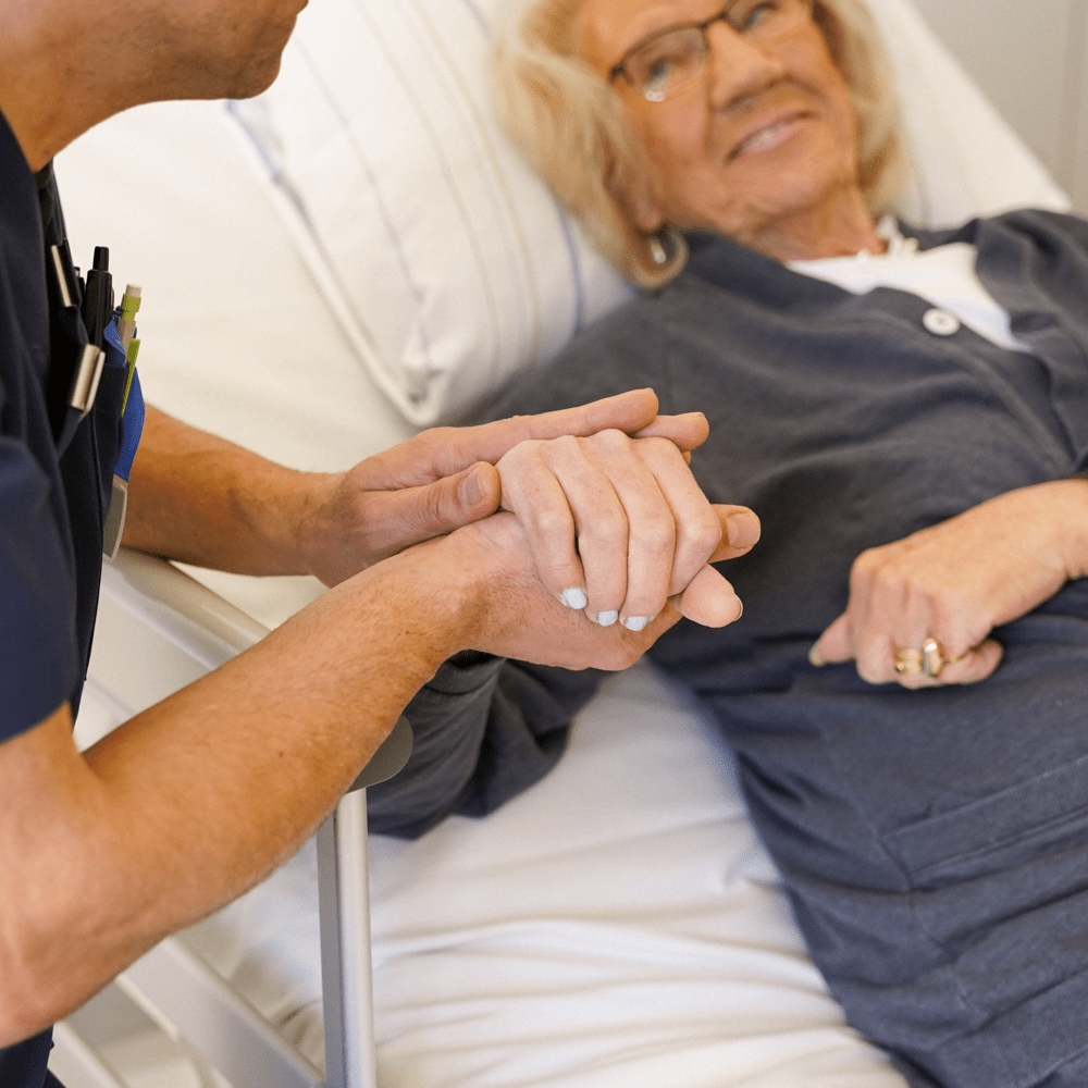 Vårdpersonal håller handen med patient som ligger i säng